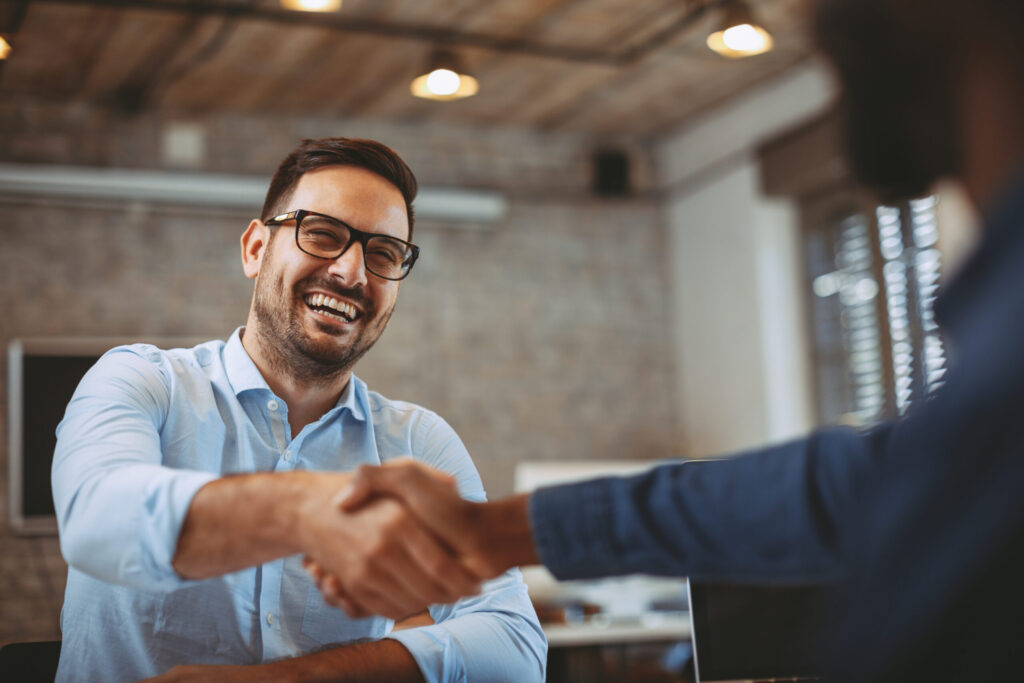 A smiling businessman with glasses shakes hands with another person in a modern office setting, symbolizing a successful deal or partnership.