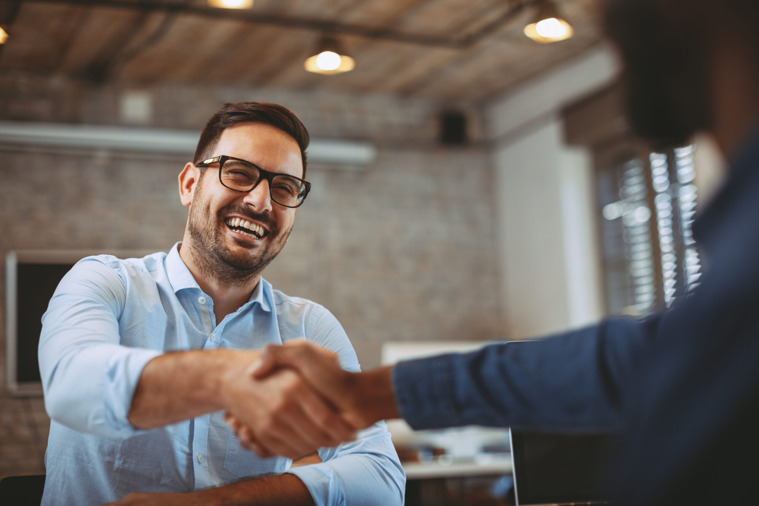 A smiling businessman with glasses shakes hands with another person in a modern office setting, symbolizing a successful deal or partnership.