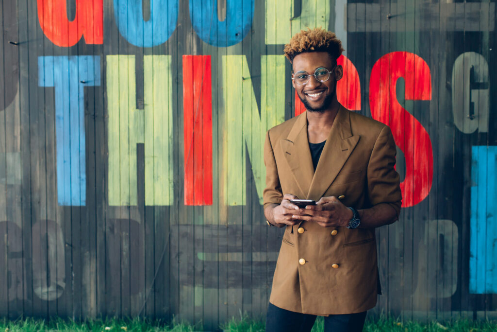 A smiling Black man with curly hair and glasses, wearing a brown double-breasted blazer, holds a smartphone while standing in front of a wooden wall with large, colorful graffiti letters.