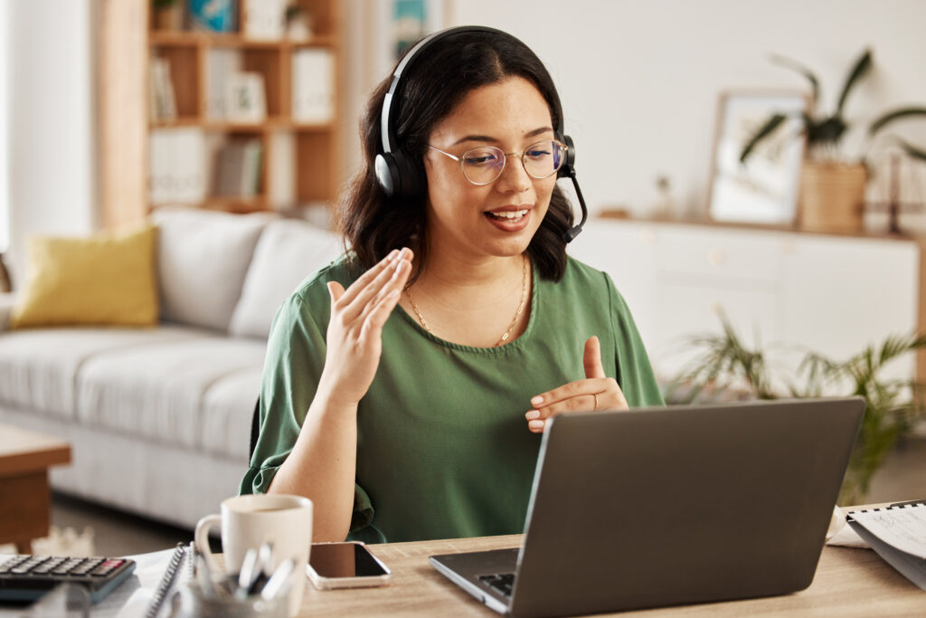 A woman with glasses and a headset is actively engaged in a video call, gesturing with her hands while looking at her laptop screen in a home office setting.