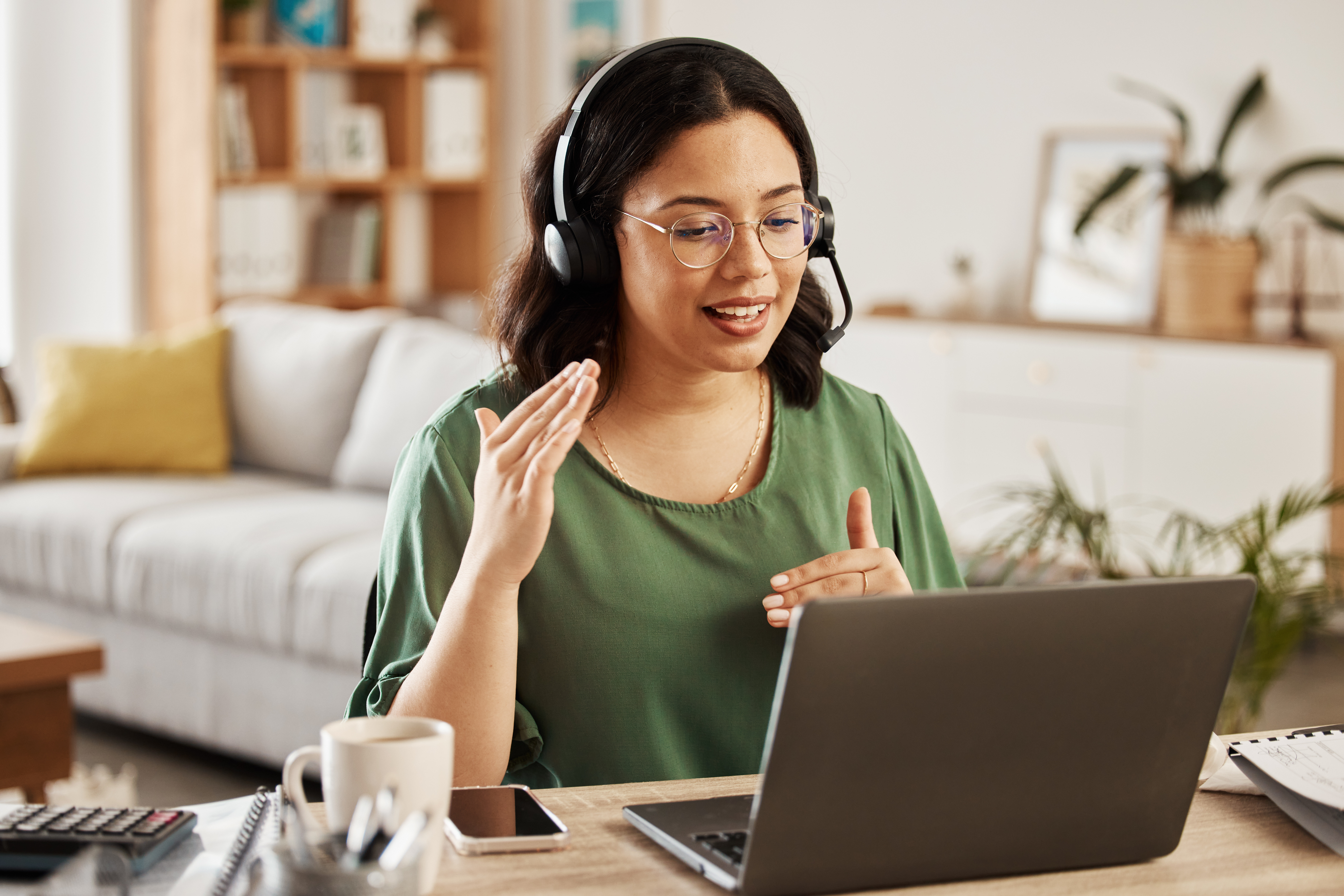A woman with glasses and a headset is actively engaged in a video call, gesturing with her hands while looking at her laptop screen in a home office setting.