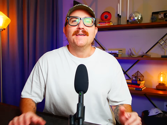 A man with a mustache and glasses wearing a white t-shirt and camo hat, speaking into a podcast microphone in a studio with purple lighting.