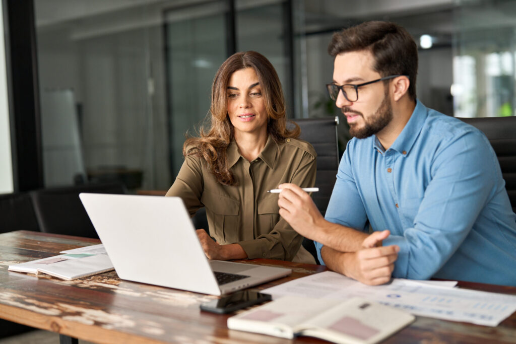 A man and a woman in a modern office setting, looking at a laptop on a wooden desk, discussing business. The man is wearing glasses and a blue shirt, the woman a green shirt.