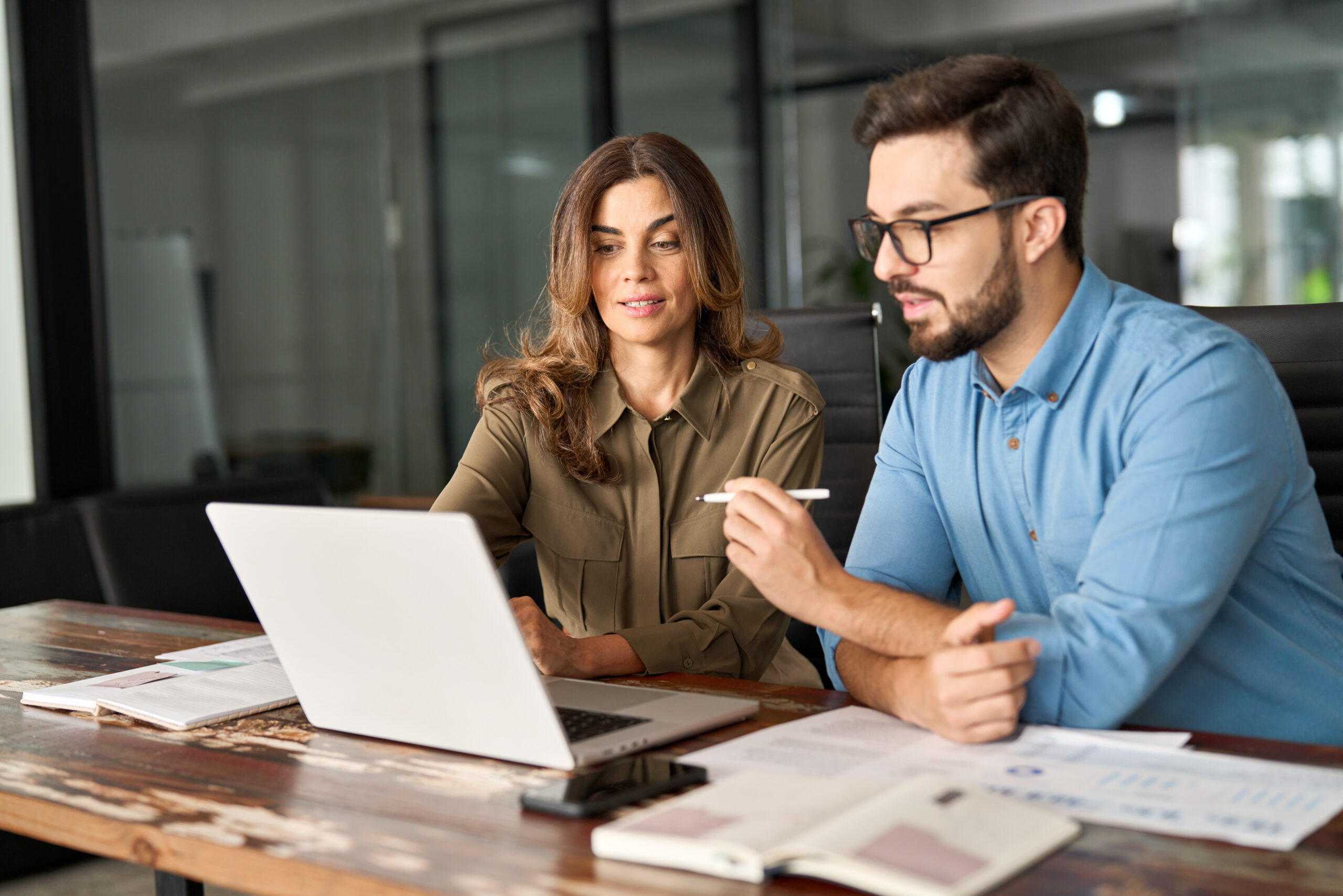 A man and a woman in a modern office setting, looking at a laptop on a wooden desk, discussing business. The man is wearing glasses and a blue shirt, the woman a green shirt.