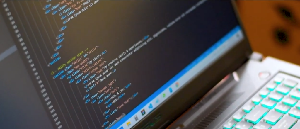 Close-up of a laptop screen displaying HTML and CSS code with line numbers, next to a white keyboard with glowing cyan backlighting.