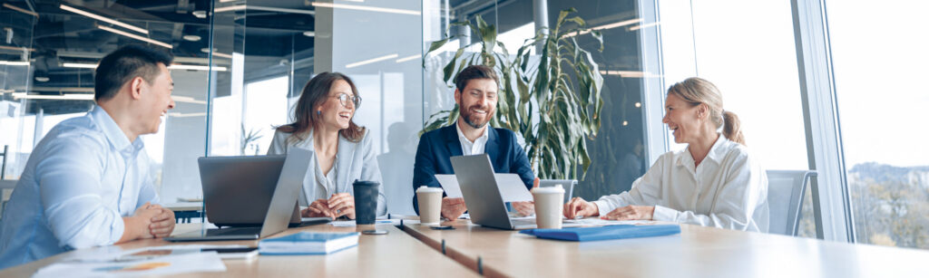 A diverse group of four professionals, two men and two women, smiling and engaged in a meeting around a large table in a bright, modern office with large windows.