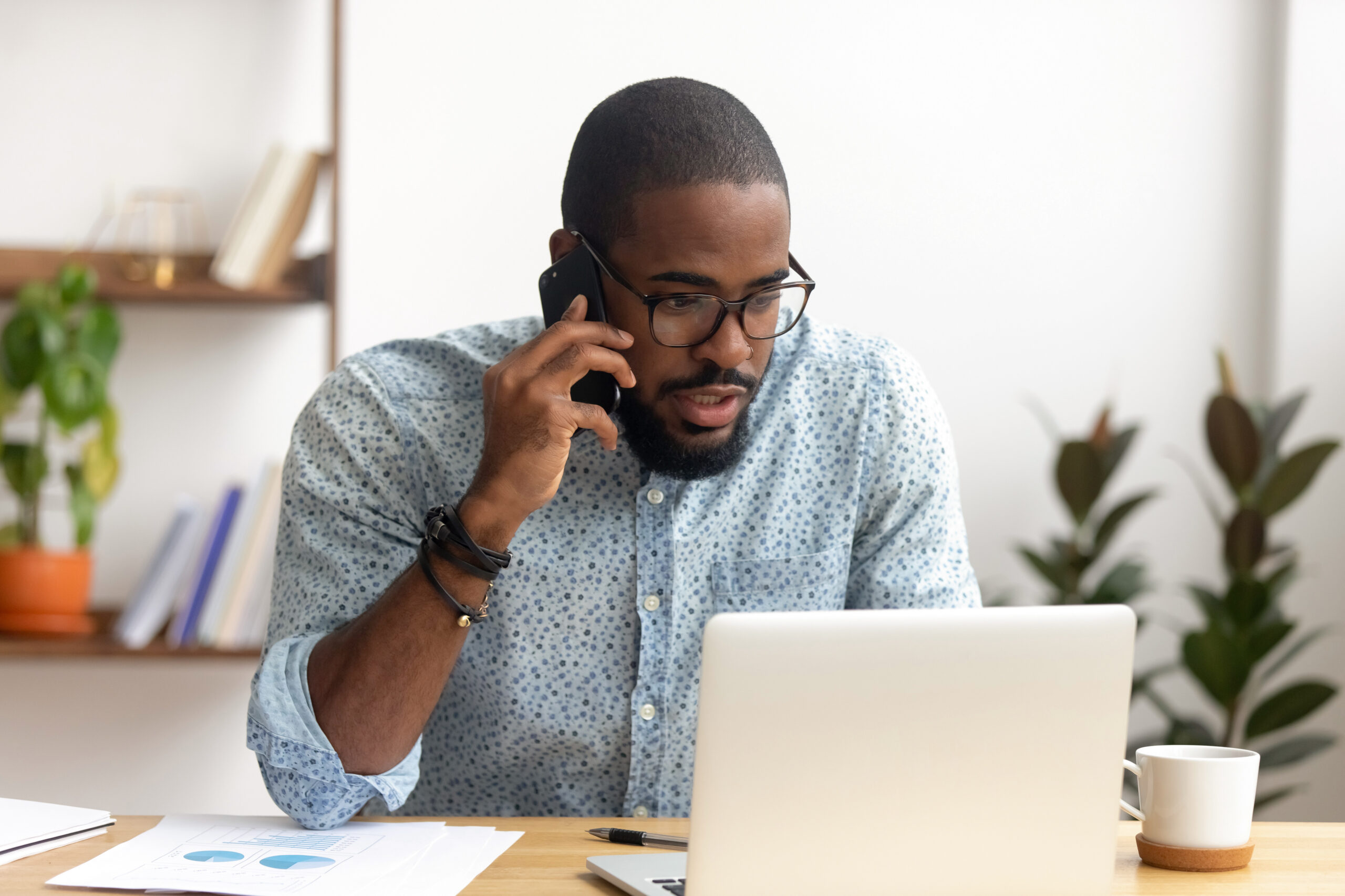 A Black man wearing glasses and a patterned shirt is on a phone call while looking at his laptop, with charts and a coffee mug on his desk.