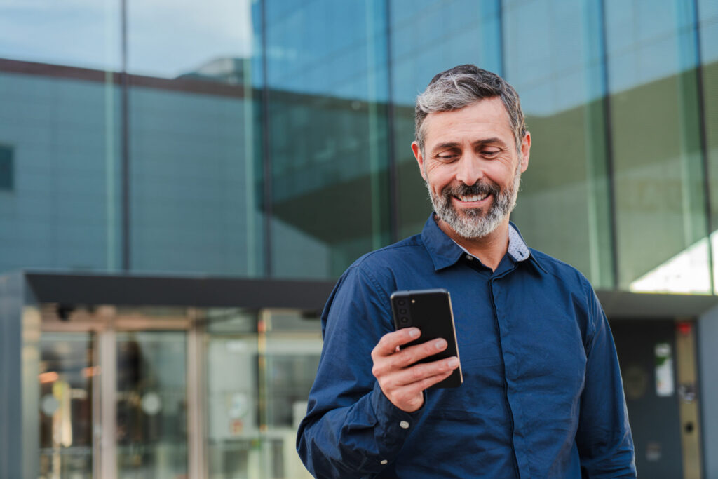 A middle-aged man with a beard and graying hair smiles as he looks at his smartphone, standing in front of a contemporary glass building.