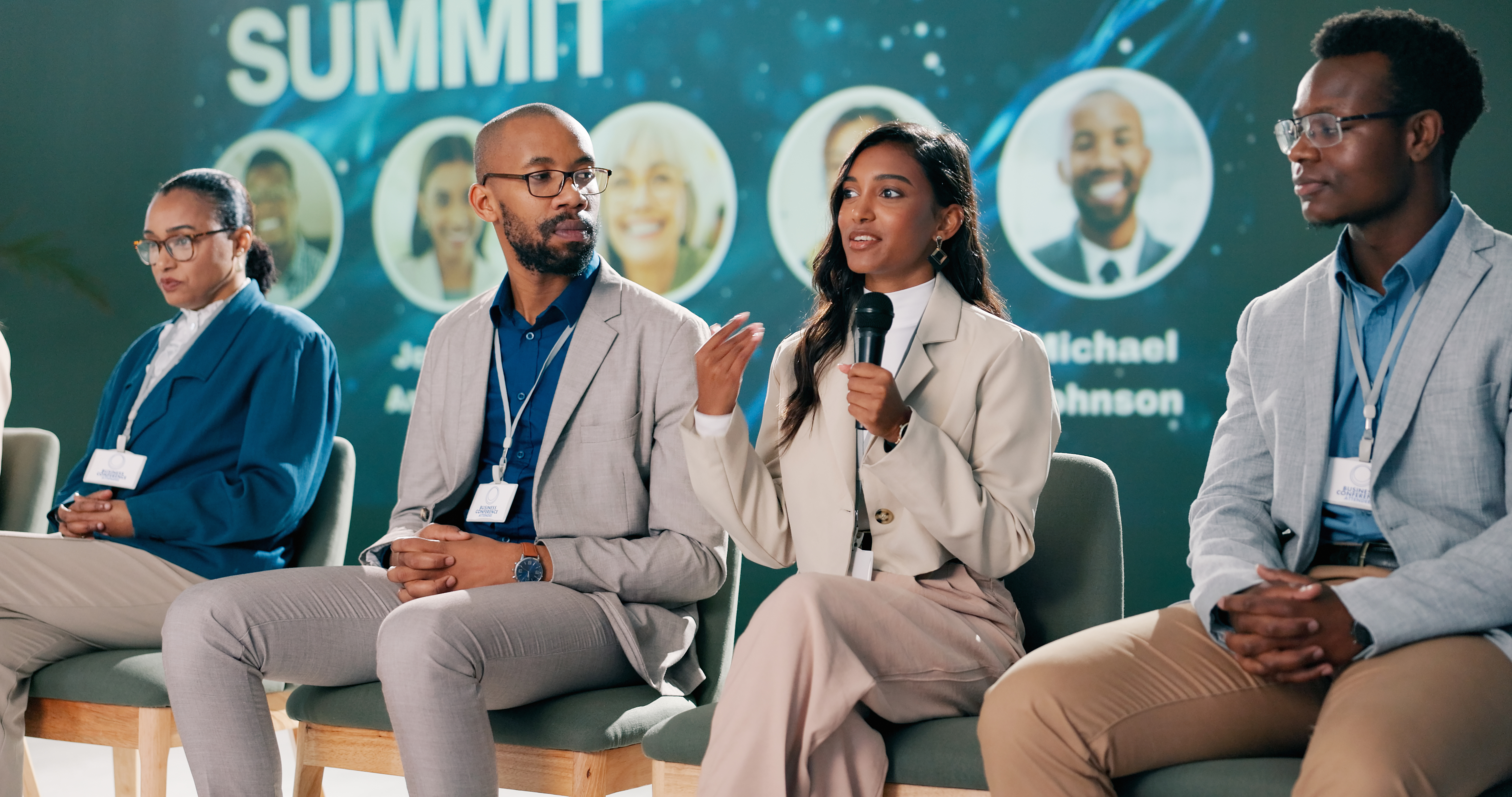 Four diverse professionals, including two women and two men, seated on a stage at a business summit, with one woman speaking into a microphone.
