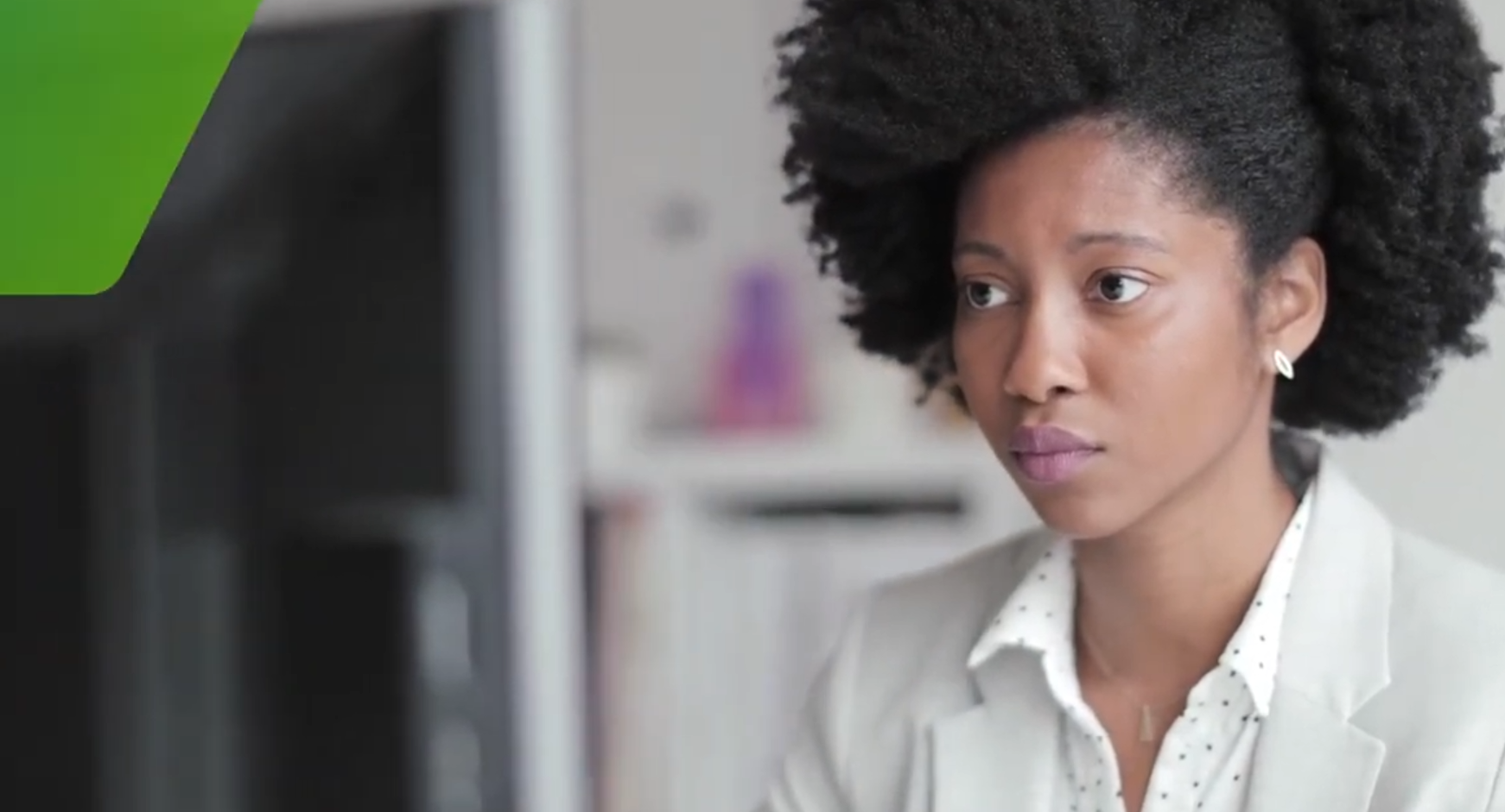 Close-up of a professional Black woman with natural afro hair wearing a light grey blazer and polka dot shirt, looking intently at a computer screen in an office setting.