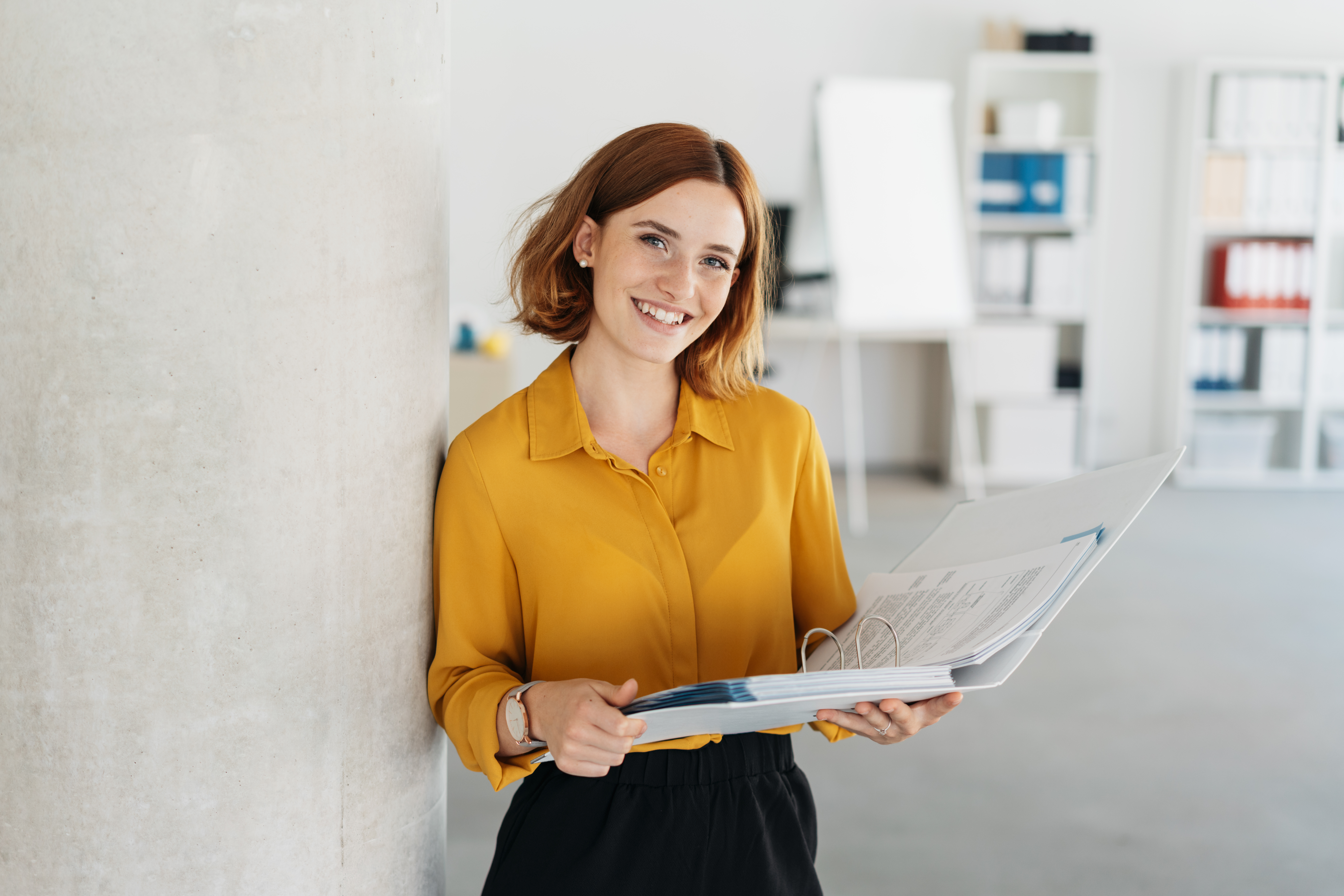 A cheerful young businesswoman with red hair and freckles smiles while holding an open binder filled with documents, leaning against a pillar in a bright office.